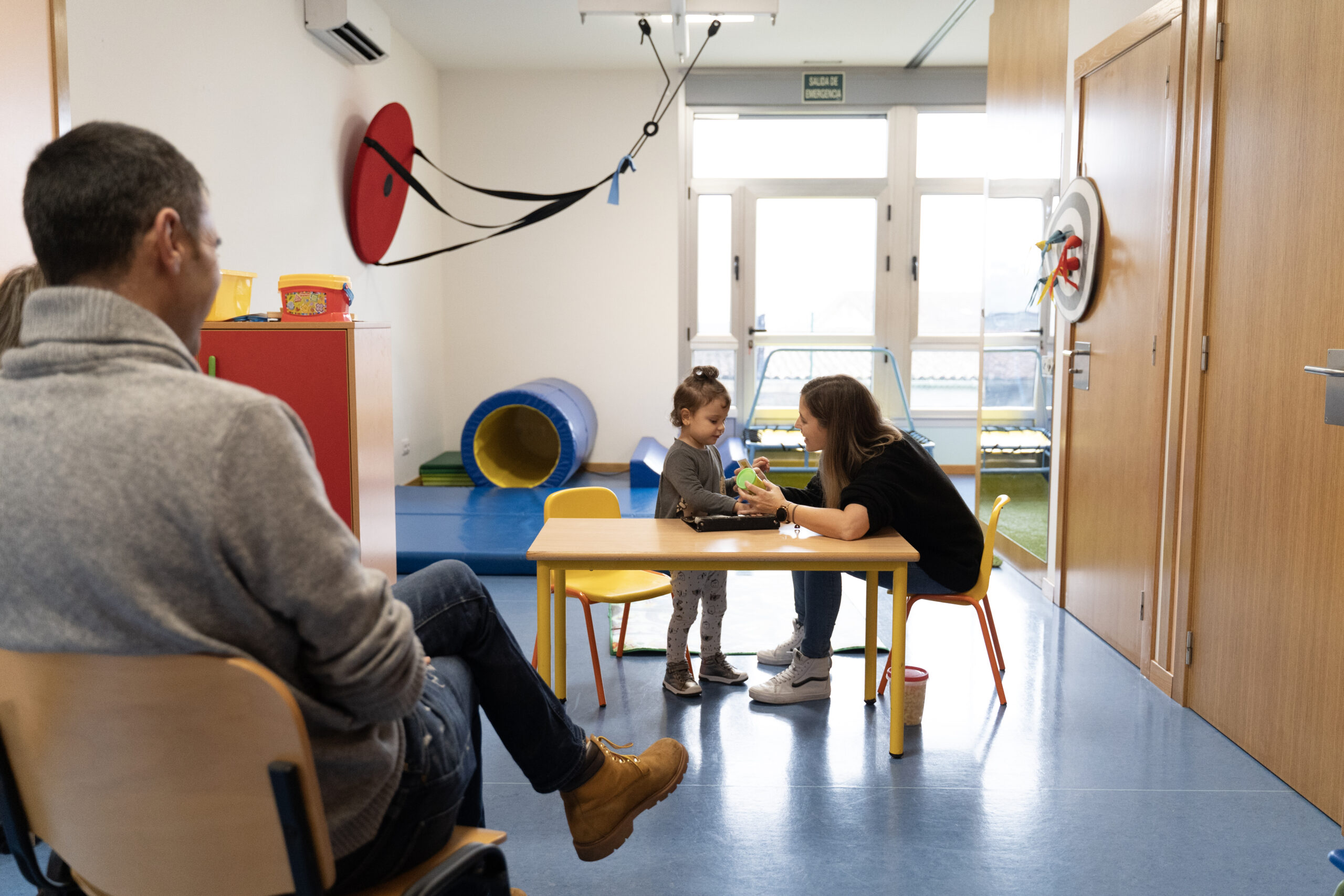 Un padre observa una sesión de intervención de niña pequeña y mujer terapeuta en una sala con una mesa.